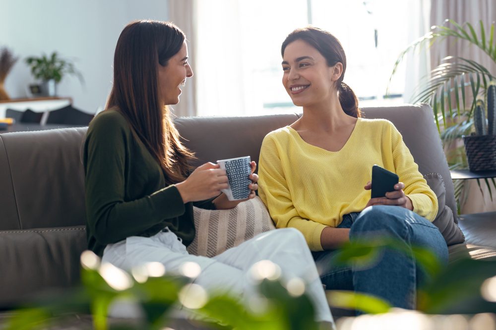 Shot of two smiling young women talking while drinking coffee sitting on couch in the living room at home.