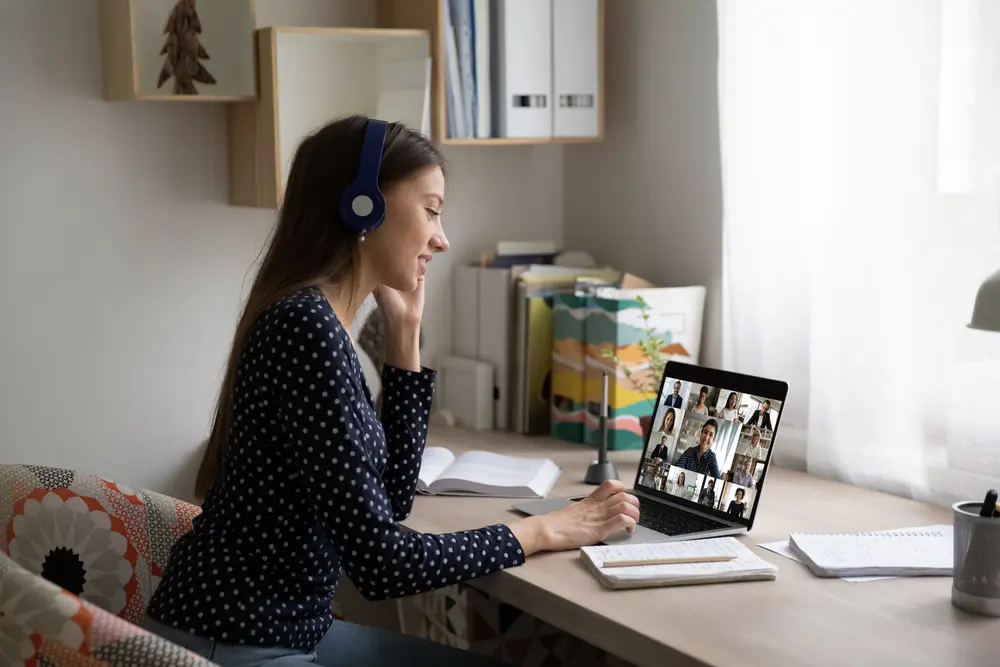 Smiling Caucasian female employee in headphones sit at desk at home talk on video call with colleagues, young woman have online webcam conversation virtual event with diverse multiethnic coworkers