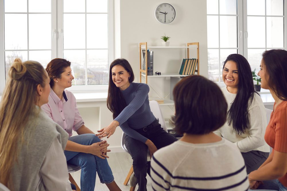 Happy positive smiling female coach, therapist or business team manager supporting and motivating young women in group therapy session or corporate staff training meeting at work